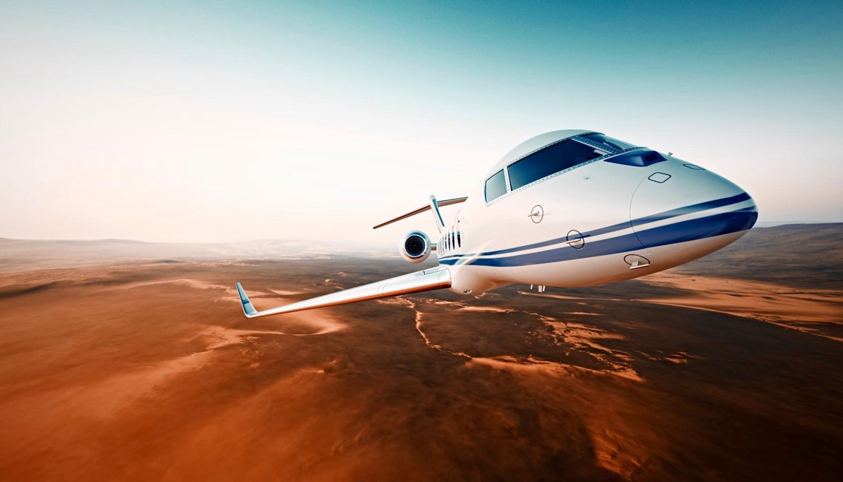 A white and blue business jet in cruise flight above red desert terrain seen from above and ahead
