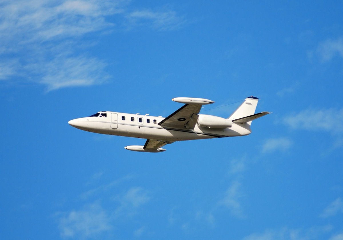 A small white business jet with wingtip tanks in flight against a blue sky with light cloud