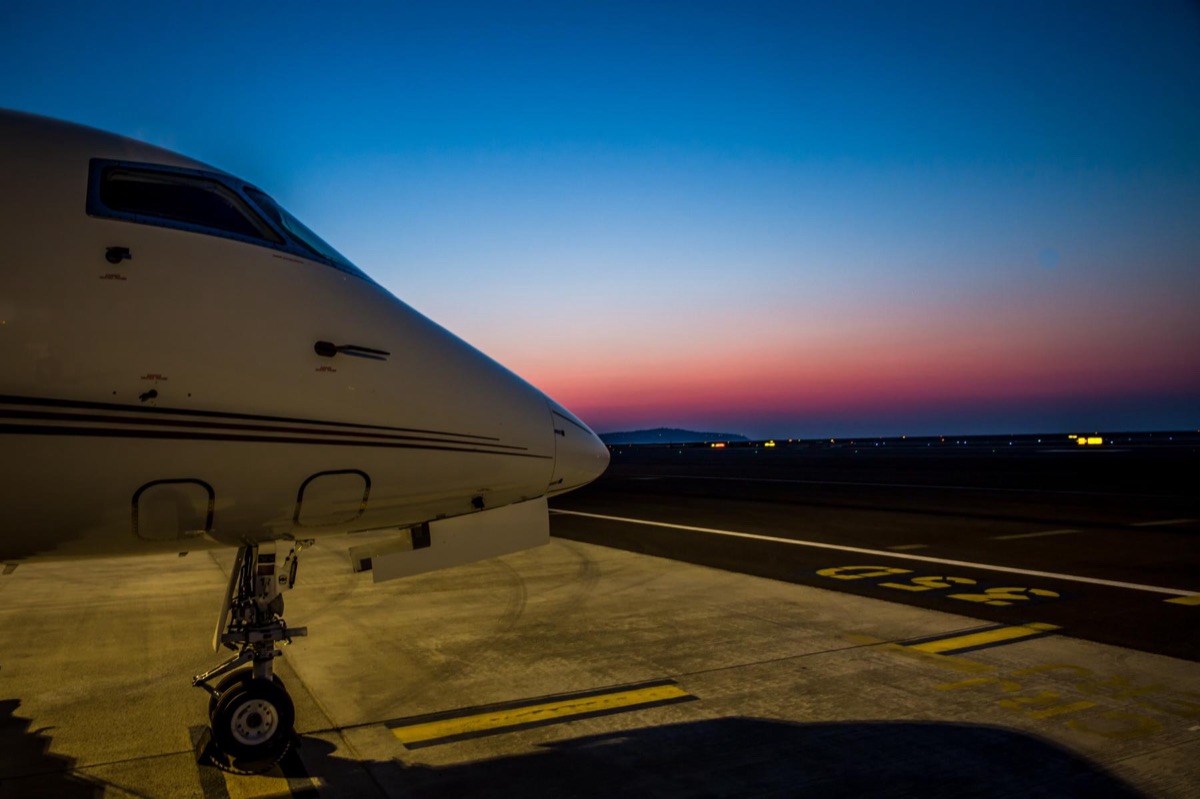 The forward fuselage and cockpit of a business jet in silhouette on the ramp at dusk, with runway edge lights and a pink and blue horizon behind