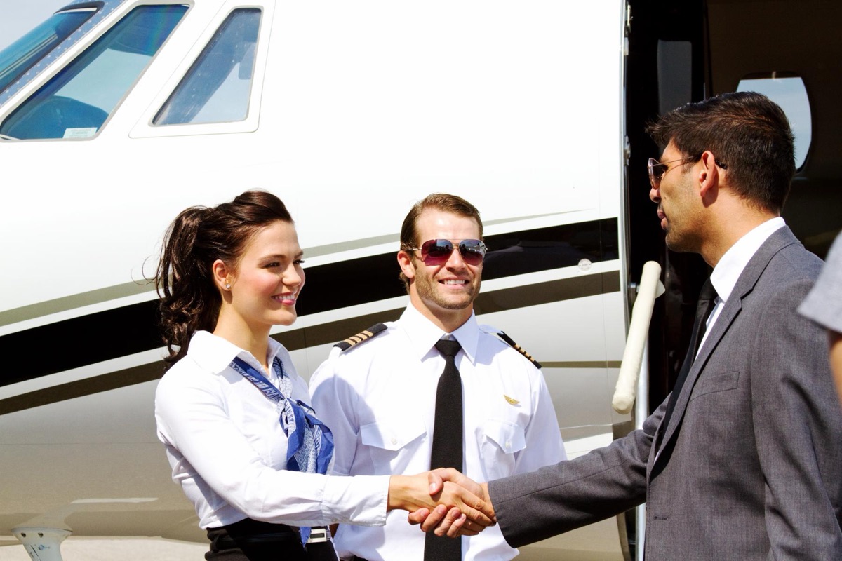 A cabin attendant and a uniformed pilot greeting a passenger in a suit with a handshake beside the open door of a private jet