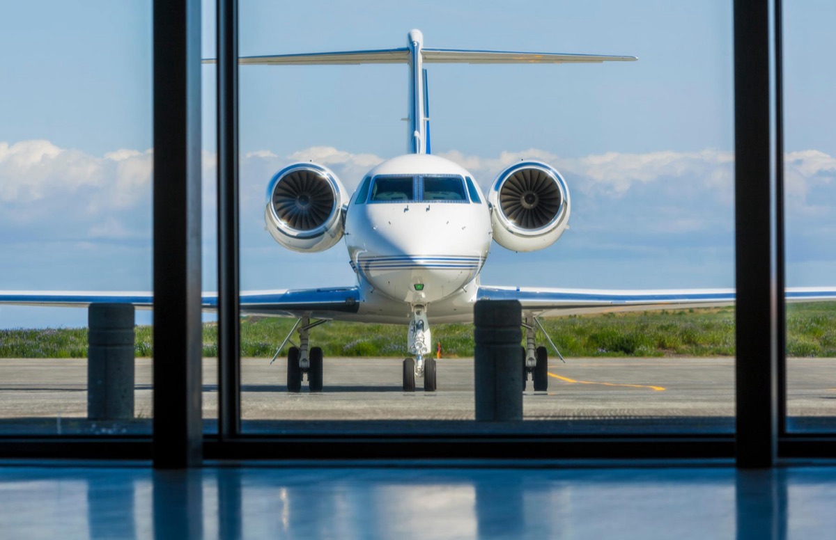A large business jet parked on the ramp seen through the floor to ceiling glass of a private terminal lounge